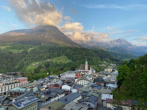 Blick auf eine Berglandschaft mit einem kleinen Ort im Vordergrund und Wolken am Himmel.