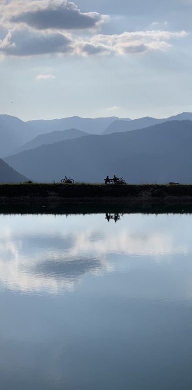 Ausblick bei einer E-Bike Tour im Nationalpark Berchtesgaden Zwei Radfahrer sitzen am Ufer eines ruhigen Speicherteichs im Nationalpark Berchtesgaden, umgeben von Bergen und Wolken.